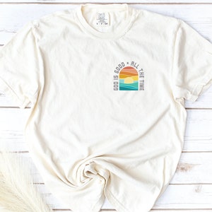 May include: A cream-colored t-shirt with a retro-style graphic design that says "God is good all the time". The graphic features a sunset with a rainbow and ocean waves.