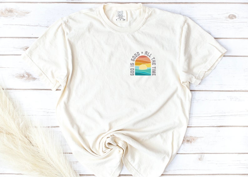 May include: A cream-colored t-shirt with a retro-style graphic design that says "God is good all the time". The graphic features a sunset with a rainbow and ocean waves.