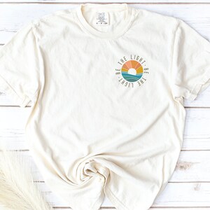 May include: A white t-shirt with a circular graphic design featuring a sunset and the text "Be the light. Be the light."