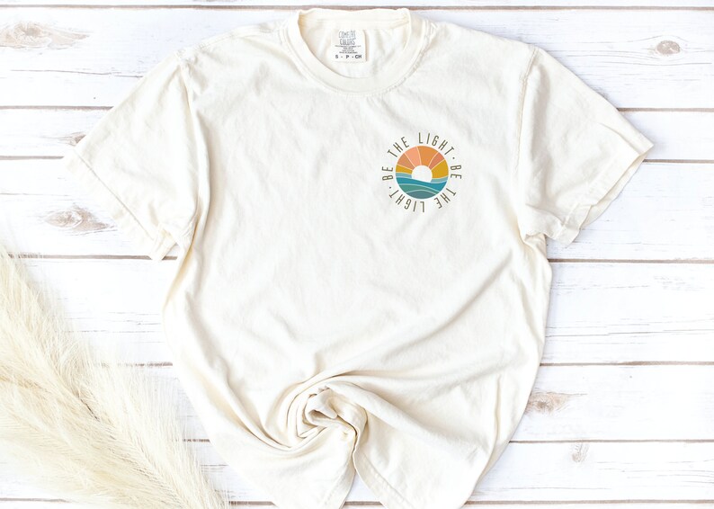 May include: A white t-shirt with a circular graphic design featuring a sunset and the text "Be the light. Be the light."