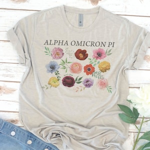Op de afbeelding: Een lichtgrijs T-shirt met een bloemenprint en de tekst "ALPHA OMICRON PI" gedrukt op de voorkant.