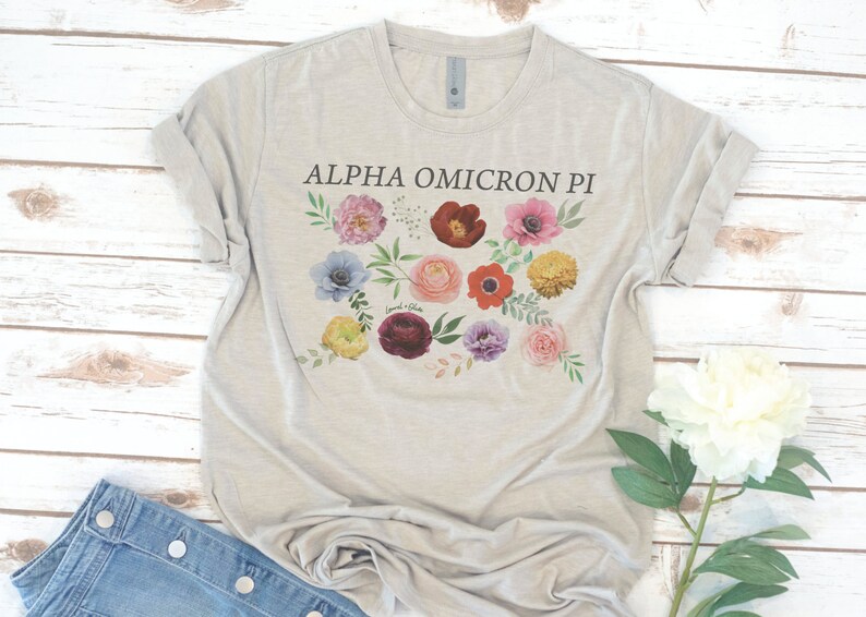 Op de afbeelding: Een lichtgrijs T-shirt met een bloemenprint en de tekst "ALPHA OMICRON PI" gedrukt op de voorkant.