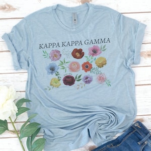 Op de afbeelding: Een lichtblauw T-shirt met de tekst "Kappa Kappa Gamma" in zwart gedrukt. Het shirt heeft een bloemenmotief met verschillende gekleurde bloemen en bladeren.