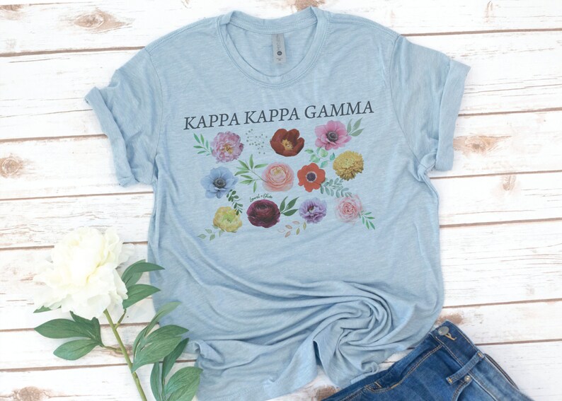 Op de afbeelding: Een lichtblauw T-shirt met de tekst "Kappa Kappa Gamma" in zwart gedrukt. Het shirt heeft een bloemenmotief met verschillende gekleurde bloemen en bladeren.