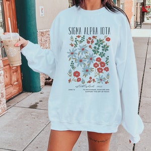Può includere: Felpa bianca con un design floreale e il testo "SIGMA ALPHA IOTA" e "established 1903" con il testo "JUNE 12 TO ENCOURAGE, NURTURE AND SUPPORT THE ART OF MUSIC" sotto.
