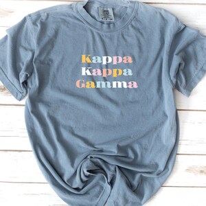 Pu&ograve; includere: Una maglietta blu chiaro con il testo "Kappa Kappa Gamma" in colori pastello.