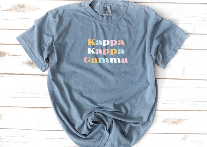 Pu&ograve; includere: Una maglietta blu chiaro con il testo "Kappa Kappa Gamma" in colori pastello.