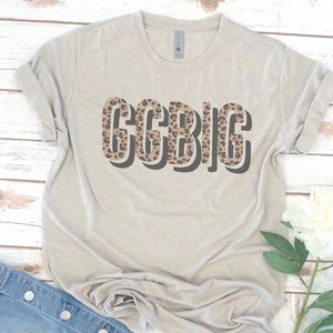 May include: A light gray t-shirt with a leopard print design that says "GCBIG" in bold, black letters.
