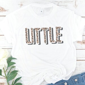May include: White t-shirt with the word "LITTLE" printed in black outlined letters. The letters are filled with a brown and black leopard print pattern.