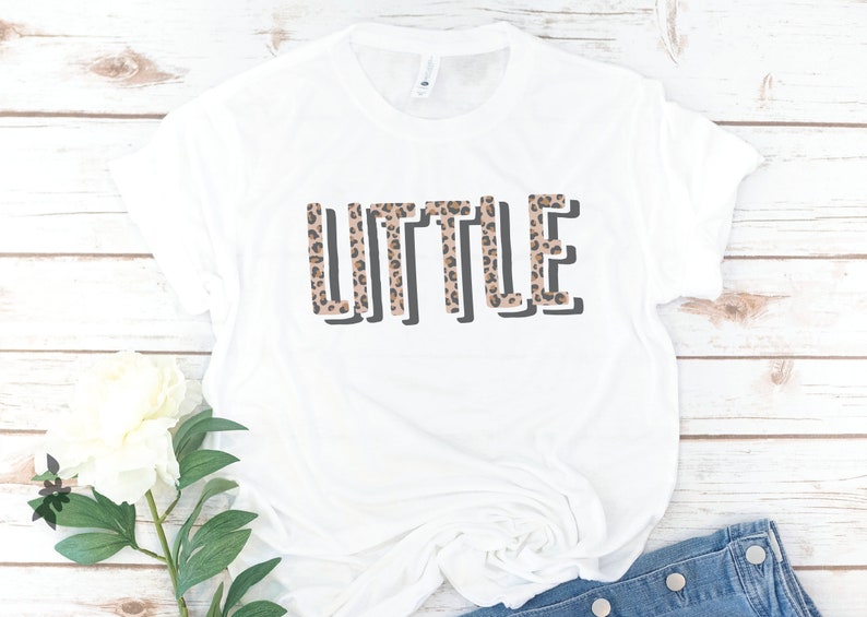 May include: White t-shirt with the word "LITTLE" printed in black outlined letters. The letters are filled with a brown and black leopard print pattern.