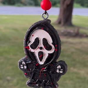 May include: A hanging ornament shaped like a hooded figure with a white mask, black glittery robe, and red paint splatters. The figure has a silver metal eyelet at the top, attached to a black string with one black and one red spherical bead.