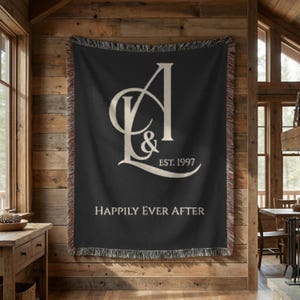 Puede incluir: Una manta tejida de color negro con bordes deshilachados que presenta un gran monograma en color crema de las letras A y L con un ampersand, el texto EST. 1997 y la frase HAPPILY EVER AFTER impresa en una fuente serif en la parte inferior.