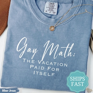Puede incluir: Una camiseta de manga corta color azul jean con cuello redondo. La prenda presenta un texto en blanco que dice "Gay Math: THE VACATION PAID FOR ITSELF". Un collar de tono dorado con un pequeño colgante circular descansa sobre el cuello.
