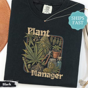Puede incluir: Una camiseta negra con un estampado gráfico de plantas de cannabis, herramientas de jardinería, un frasco de vidrio y un atomizador. El texto "Plant Manager" está impreso en color beige sobre y bajo la ilustración. Tiene cuello redondo.