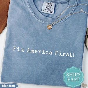 Puede incluir: Una camiseta de color azul vaquero con el texto blanco Fix America First! impreso en la parte delantera. La prenda cuenta con cuello redondo, una etiqueta de la marca Comfort Colors y una fina cadena dorada con un pequeño colgante circular.
