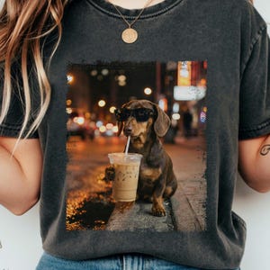 Op de afbeelding: Donkergrijs T-shirt met een afbeelding van een teckel met een zonnebril en een ijskoffie. De hond zit op een stoeprand met een vage stadsachtergrond. De afbeelding heeft een vintage, versleten uitstraling.