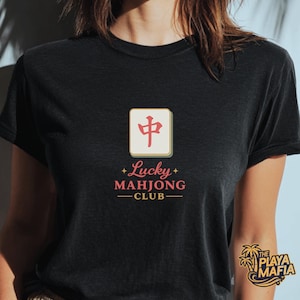 May include: Black t-shirt featuring a Mahjong tile graphic with the text "Lucky Mahjong Club" in pink and gold. The design includes a red Chinese character. The Playa Mafia logo is visible on the lower right.