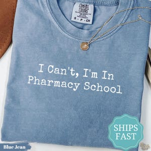 May include: A blue jean colored t-shirt with a crew neckline and a white graphic print that reads I Can't, I'm In Pharmacy School. The shirt features a Comfort Colors brand label at the neck and a thin gold chain necklace resting on the fabric.