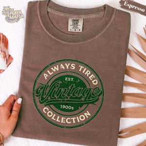 May include: Espresso-colored t-shirt with a green and white circular graphic that reads "ALWAYS TIRED Vintage Collection 1900s." The shirt has a crew neck and short sleeves. The brand tag is visible at the neckline.