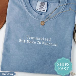 May include: A blue jean colored t-shirt with a crew neckline and a white embroidered text that reads Traumatized But Make It Fashion. A thin gold chain necklace with a small circular pendant rests on the fabric near the collar of the shirt.