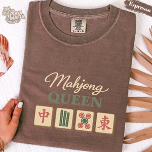 May include: Espresso-colored t-shirt with the words "Mahjong QUEEN" in a stylish font. Below the text are four Mahjong tiles. The shirt has a crew neck and a Comfort Colors tag.