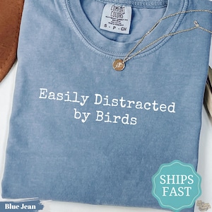 May include: A blue jean colored t-shirt with a crew neckline and a white graphic print that reads "Easily Distracted by Birds". A gold-toned chain necklace with a small circular pendant rests on the fabric near the collar of the shirt.