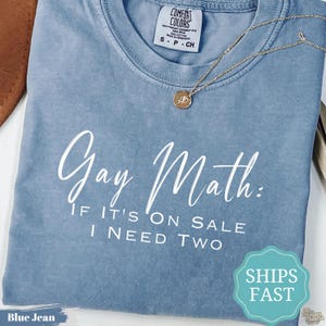 Puede incluir: Una camiseta de manga corta color azul jean con cuello redondo. La prenda presenta un texto en blanco que dice, Gay Math: IF IT'S ON SALE I NEED TWO. Un collar de tono dorado con un pequeño colgante circular descansa sobre el cuello.