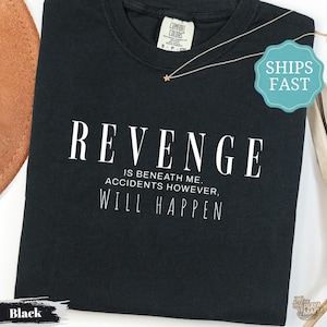 以下が含まれることがあります： REVENGE IS BENEATH ME. ACCIDENTS HOWEVER, WILL HAPPENという白い文字がプリントされた黒いクルーネックTシャツ。襟の内側にはComfort Colorsのブランドラベルがあり、生地の上には細いゴールドトーンのネックレスが置かれています。