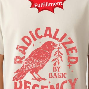 Puede incluir: Camiseta color crema con un gráfico rojo desgastado de un cuervo sosteniendo una ramita, rodeado por las palabras "RADICALIZED BY BASIC DECENCY." Un gráfico rojo en forma de estrella en la parte superior dice "Fast Order Fulfillment."