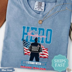 May include: A light blue t-shirt featuring a graphic of a person in a military uniform holding a sign that reads IMPEACH CONVICT REMOVE in front of an American flag. The text HERO, American, and I STAND WITH MAJOR WATSON are printed on the shirt.