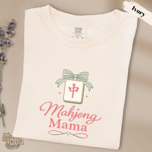 Pode incluir: Camiseta cor marfim com um design Mahjong Mama. O design apresenta uma peça de Mahjong com um caractere vermelho, um laço verde e as palavras "Mahjong Mama" em script rosa. A camisa está dobrada.