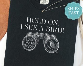 May include: A black t-shirt featuring the white text HOLD ON, I SEE A BIRD! above a graphic of binoculars. The left lens shows a tree branch with leaves, and the right lens shows a small bird perched on a branch. The shirt has a crew neckline.