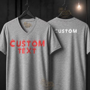 Könnte beinhalten: Zwei graue V-Ausschnitt-T-Shirts. Auf der Vorderseite eines Shirts steht "CUSTOM TEXT" in roter Flammen-Schrift. Auf der Rückseite eines Shirts steht "CUSTOM" in Weiß. Die Shirts hängen an schwarzen Kleiderbügeln.