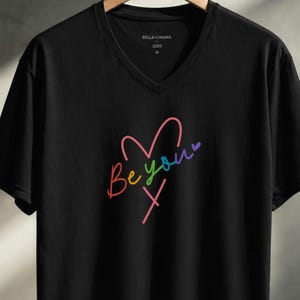 Può includere: T-shirt nero con scollo a V con un design a forma di cuore colorato e le parole "Be you" in un gradiente arcobaleno. La maglietta è di taglia M ed è realizzata in un materiale morbido.