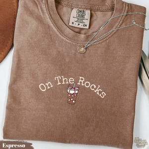 Può includere: Una t-shirt marrone con scollo rotondo e un'etichetta Comfort Colors sul colletto. Il petto presenta una scritta ricamata in bianco che recita On The Rocks sopra una piccola grafica di un caffè freddo con un fiocco rosa e una cannuccia.