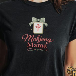 May include: Black t-shirt with a graphic design featuring a Mahjong tile with a bow and the words "Mahjong Mama" in pink script. The design includes decorative elements and is centered on the shirt.