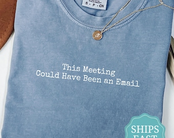May include: A blue jean colored t-shirt with a crew neckline and a white embroidered text that reads, This Meeting Could Have Been an Email. A gold chain necklace with a small circular pendant rests on the collar area of the garment.