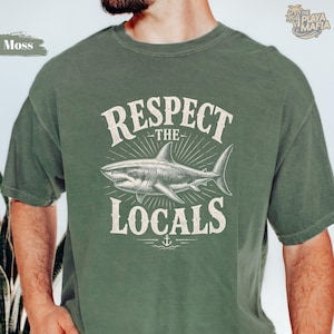 May include: A sage green short-sleeve t-shirt featuring a graphic of a shark swimming horizontally. Above the shark is the text "RESPECT" and below is "THE LOCALS" with a small anchor icon. The design is rendered in a distressed white ink.