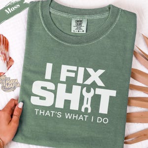 May include: A moss green t-shirt with the text "I FIX SHIT THAT'S WHAT I DO" in white. The shirt has a crew neck and short sleeves. The brand label "COMFORT COLORS" is visible on the collar.