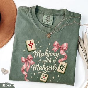 Puede incluir: Camiseta verde musgo con el texto "Mahjong with Mahgirls" e ilustraciones de fichas de mahjong y lazos rosas. La camiseta tiene cuello redondo y mangas cortas. También se ve un collar plateado con un colgante.