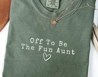 May include: A moss green crew neck t-shirt featuring the white text "Off To Be The Fun Aunt" with a small heart icon below the lettering. The shirt includes a Comfort Colors brand label at the neckline and a delicate gold-toned chain necklace.