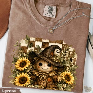 Puede incluir: Una camiseta color espresso con un gráfico de un espantapájaros con sombrero de ala ancha, rodeado de girasoles amarillos y flores blancas sobre un fondo a cuadros. La etiqueta interior del cuello dice Comfort Colors S P CH.