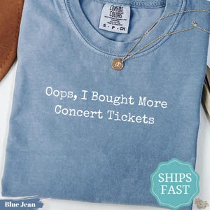 Könnte beinhalten: Ein blaues T-Shirt in Jeansoptik mit Rundhalsausschnitt und einem weißen grafischen Aufdruck mit der Aufschrift, Oops, I Bought More Concert Tickets. Das Shirt hat ein Comfort Colors Markenetikett am Hals und eine dünne goldene Halskette.