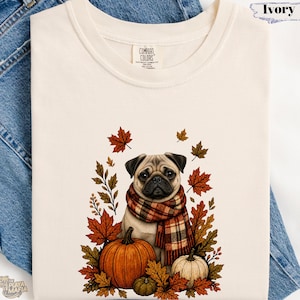May include: An ivory t-shirt featuring a graphic of a pug wearing a plaid scarf, surrounded by autumn leaves and three pumpkins. The shirt has a Comfort Colors brand label at the neck and is displayed against a background of blue denim fabric.