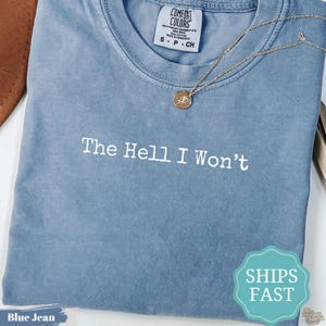 以下が含まれることがあります： 胸元に白い文字で「The Hell I Won't」とプリントされた、クルーネックの青い半袖Tシャツ。小さな円形のペンダントが付いたゴールドトーンのチェーンネックレスが、襟元付近の生地の上に置かれています。