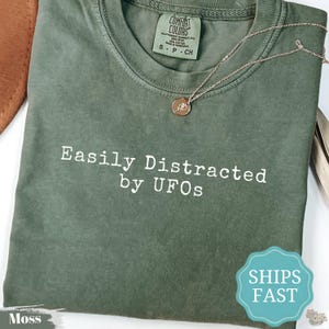 以下が含まれることがあります： モスグリーンの半袖Tシャツで、クルーネックと胸元に白い文字で「Easily Distracted by UFOs」とプリントされています。小さな円形のペンダントが付いた細いゴールドトーンのチェーンネックレスが、衣服の襟元に置かれています。