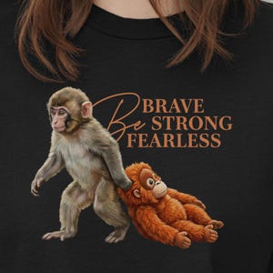 May include: Black t-shirt featuring two cartoon monkeys. One is standing, the other is lying down. The text "Be Brave Strong Fearless" is in a copper color.