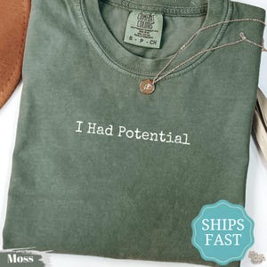 Puede incluir: Una camiseta de cuello redondo color verde musgo con el texto bordado en blanco I Had Potential centrado en el pecho. La camiseta incluye una etiqueta de la marca Comfort Colors en el cuello interior y se combina con una fina cadena dorada con colgante.