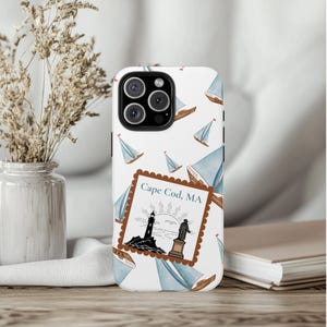 May include: White phone case featuring a nautical design with sailboats and a stamp that reads "Cape Cod, MA." The case has a lighthouse and a statue illustration. The sailboats are blue and brown.