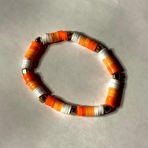 May include: A handmade bracelet featuring orange, white, and gold-colored beads. The bracelet is made of small, flat, round beads in shades of orange and white, separated by gold-colored metal beads. The bracelet is a stylish accessory.